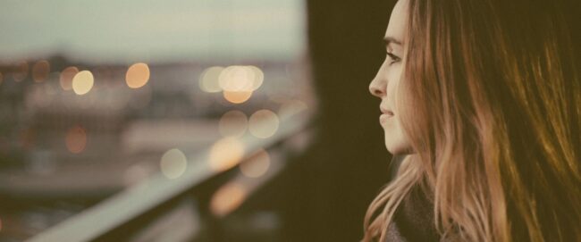 Woman gazing thoughtfully out a window with blurred city lights.