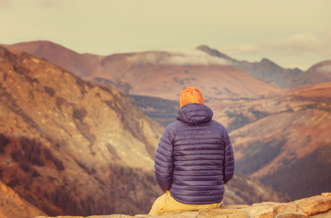 Person in a warm jacket overlooking a mountainous landscape at sunset.
